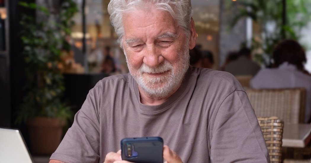 Symbolbild: Älterer Mann mit weißem Bart schaut lächelnd auf sein Smartphone, daneben ein Laptop